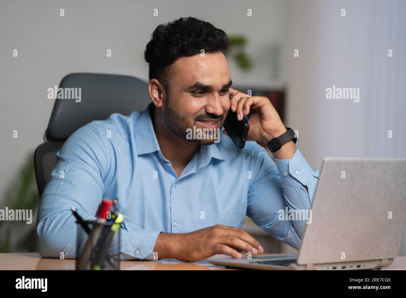 Homme d'affaires indien parlant sur le téléphone mobile tout en travaillant sur ordinateur portable au bureau - concept de conseiller financier, multi-tâches et style de vie d'entreprise Banque D'Images