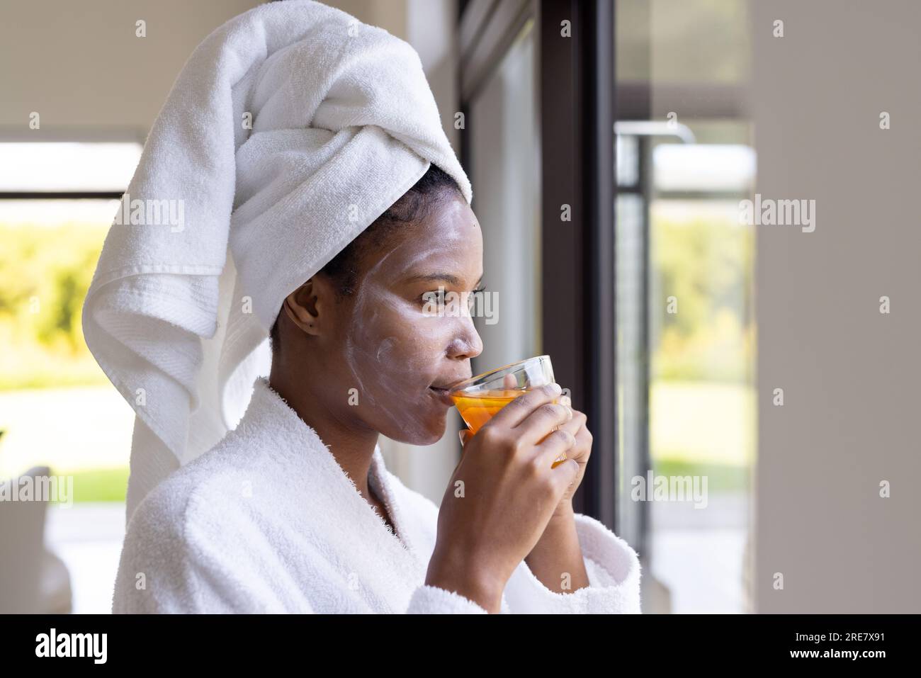 Femme afro-américaine portant une serviette et un masque facial de beauté, buvant du thé et regardant par la fenêtre Banque D'Images