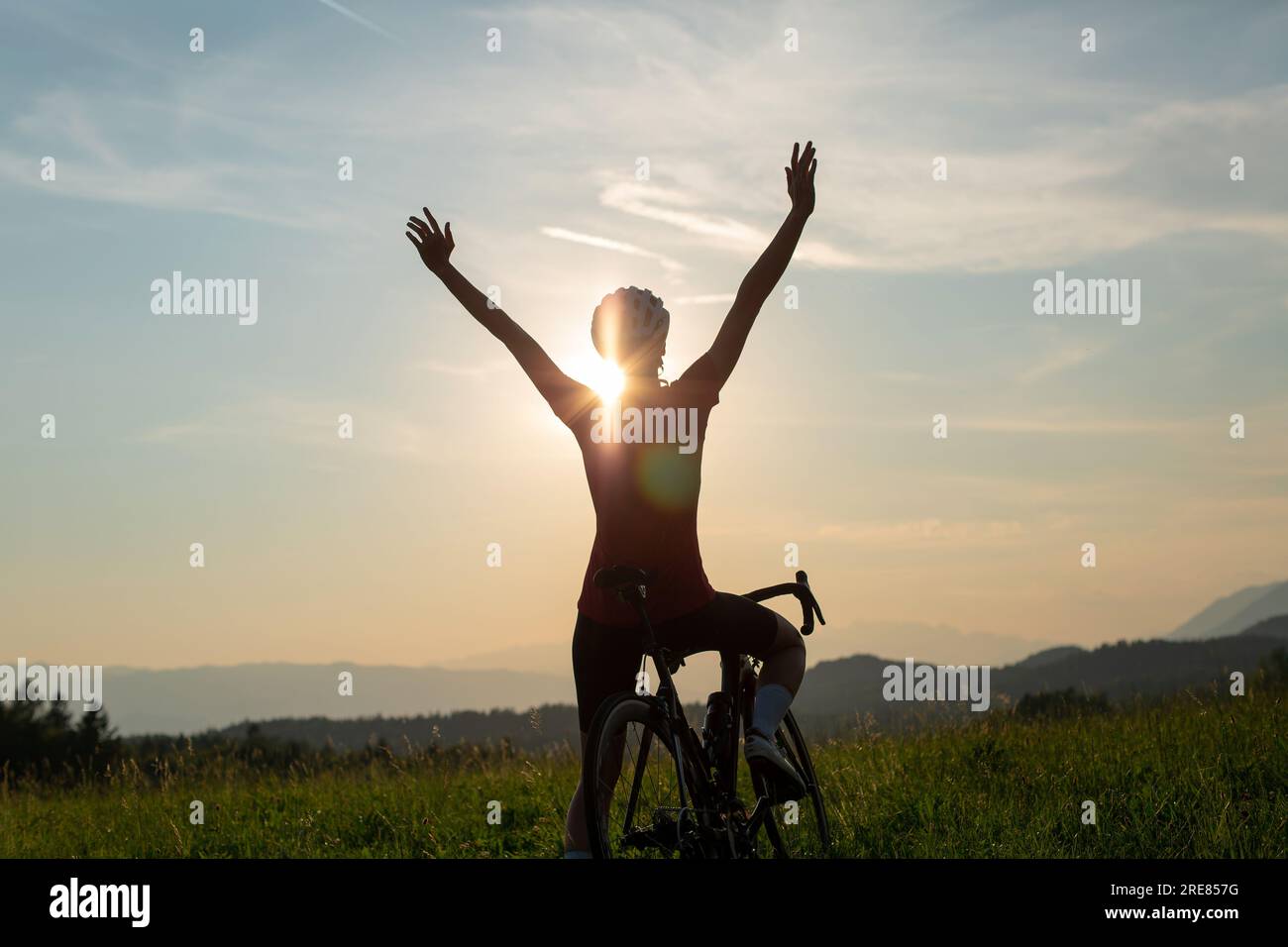 Femme sur un vélo de course s'arrêtant à une glade verte, célébrant avec les bras levés tout en regardant un incroyable panorama de montagne au coucher du soleil Banque D'Images