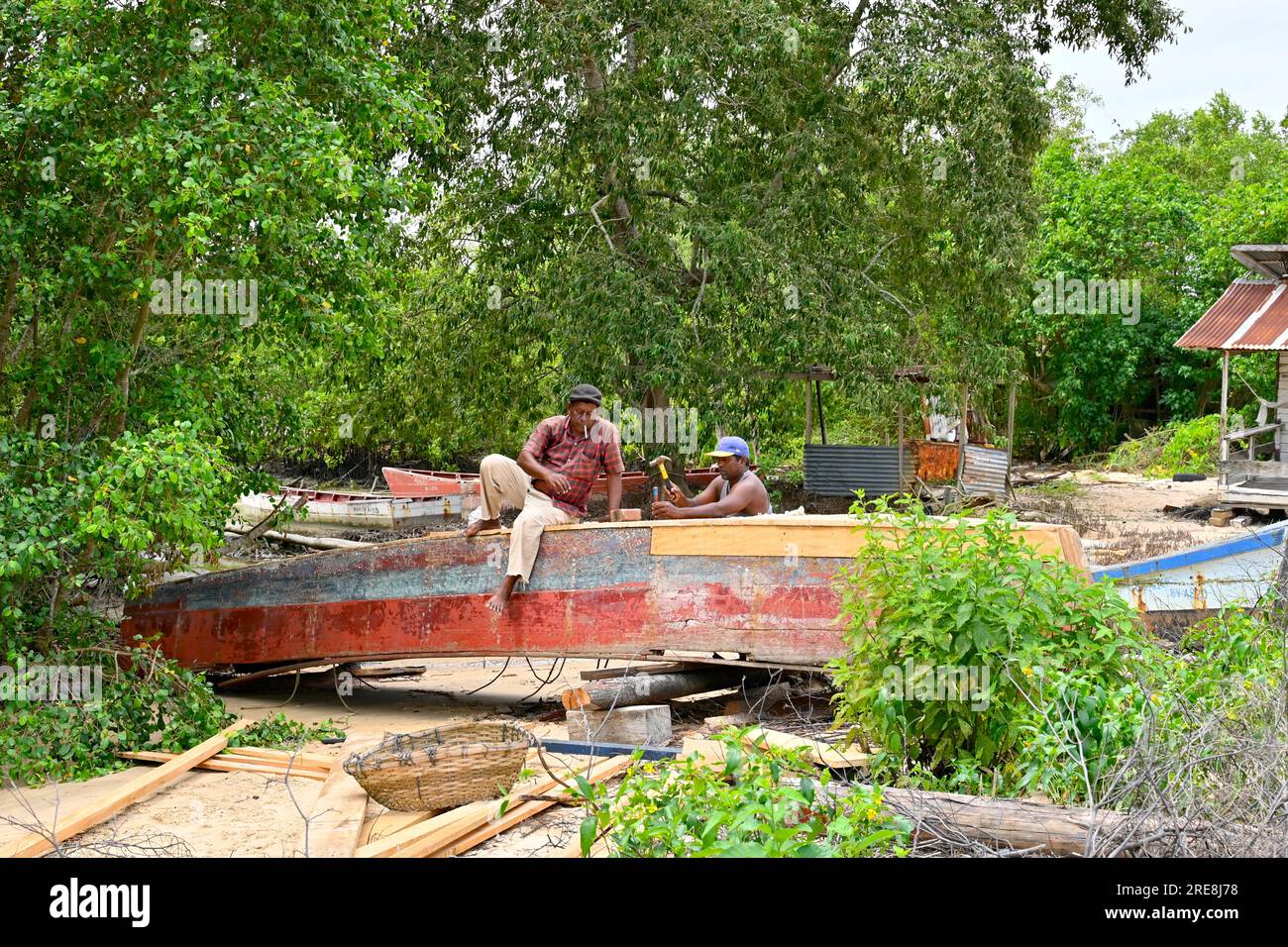 Près d'une rivière et entouré de verdure deux charpentiers du village de Ponoma au Suriname renouvellent le fond d'un bateau en bois Banque D'Images