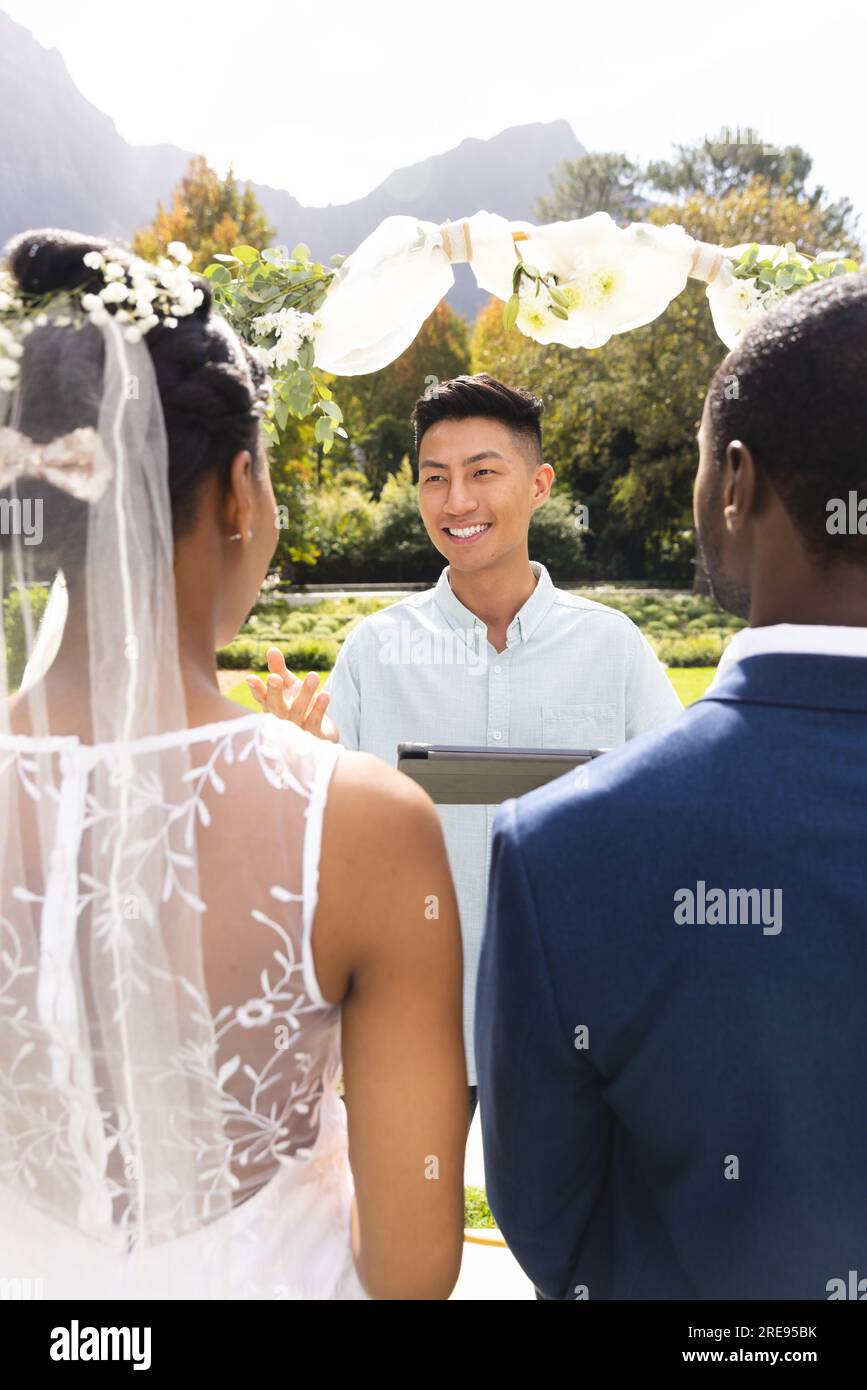 Heureux diverse officiant masculin utilisant la tablette, avec mariée et marié à la cérémonie de mariage dans le jardin ensoleillé Banque D'Images