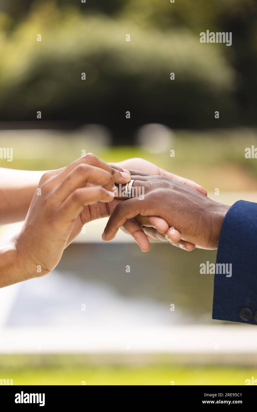 Mains de mariée afro-américaine plaçant bague sur le doigt du marié dans le jardin ensoleillé, espace de copie Banque D'Images