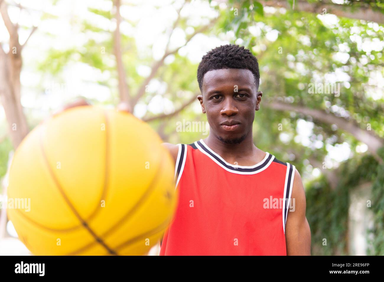 Portrait d'un jeune sportif afro-américain confiant en vêtements d'activité tenant le basket-ball tout en regardant la caméra près des arbres brouillés Banque D'Images