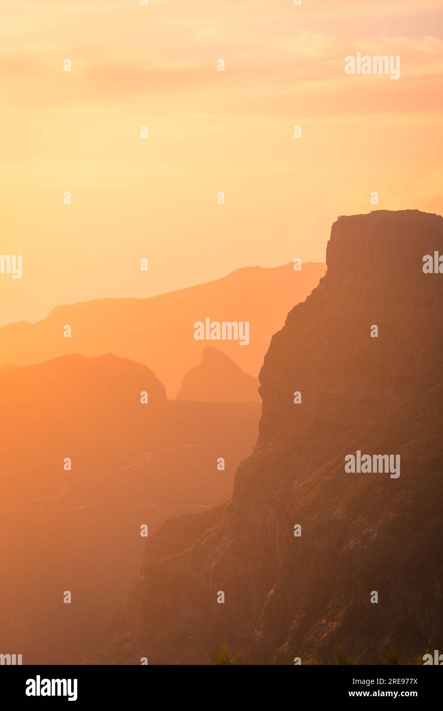 Vue pittoresque du paysage brumeux d'énormes montagnes rocheuses rugueuses avec des arbres verts sous le ciel bleu avec des nuages orange au coucher du soleil à Tenerife Espagne Banque D'Images