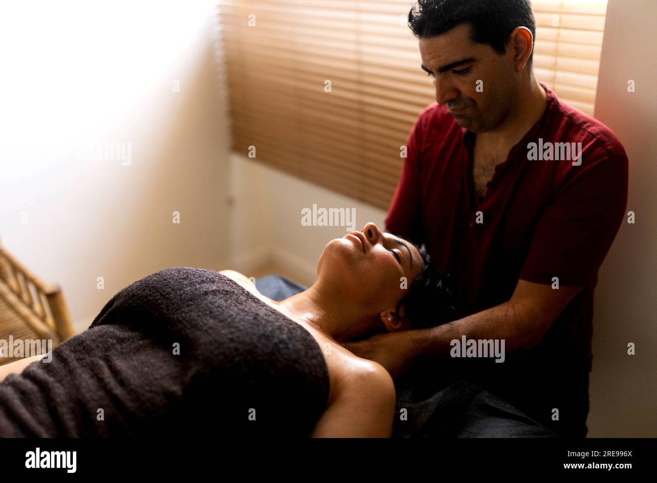 Angle élevé de masseur hispanique adulte assis près de la jeune femme hispanique couchée sur la table de massage dans le salon et traitant avec un tissu dans les mains dans la journée Banque D'Images