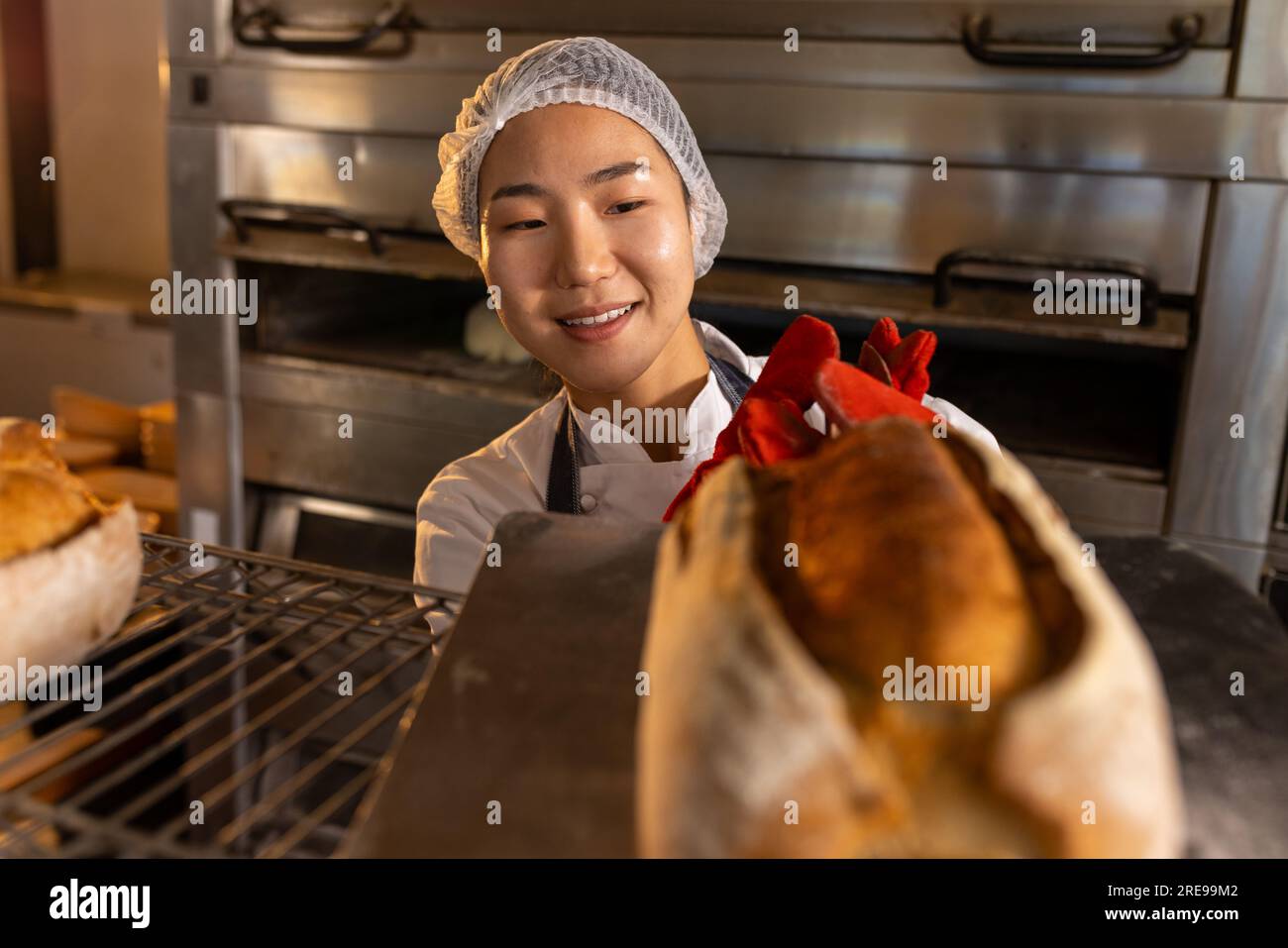 Heureuse boulangère asiatique dans la cuisine de boulangerie portant un tablier et tenant une plaque de cuisson avec du pain Banque D'Images