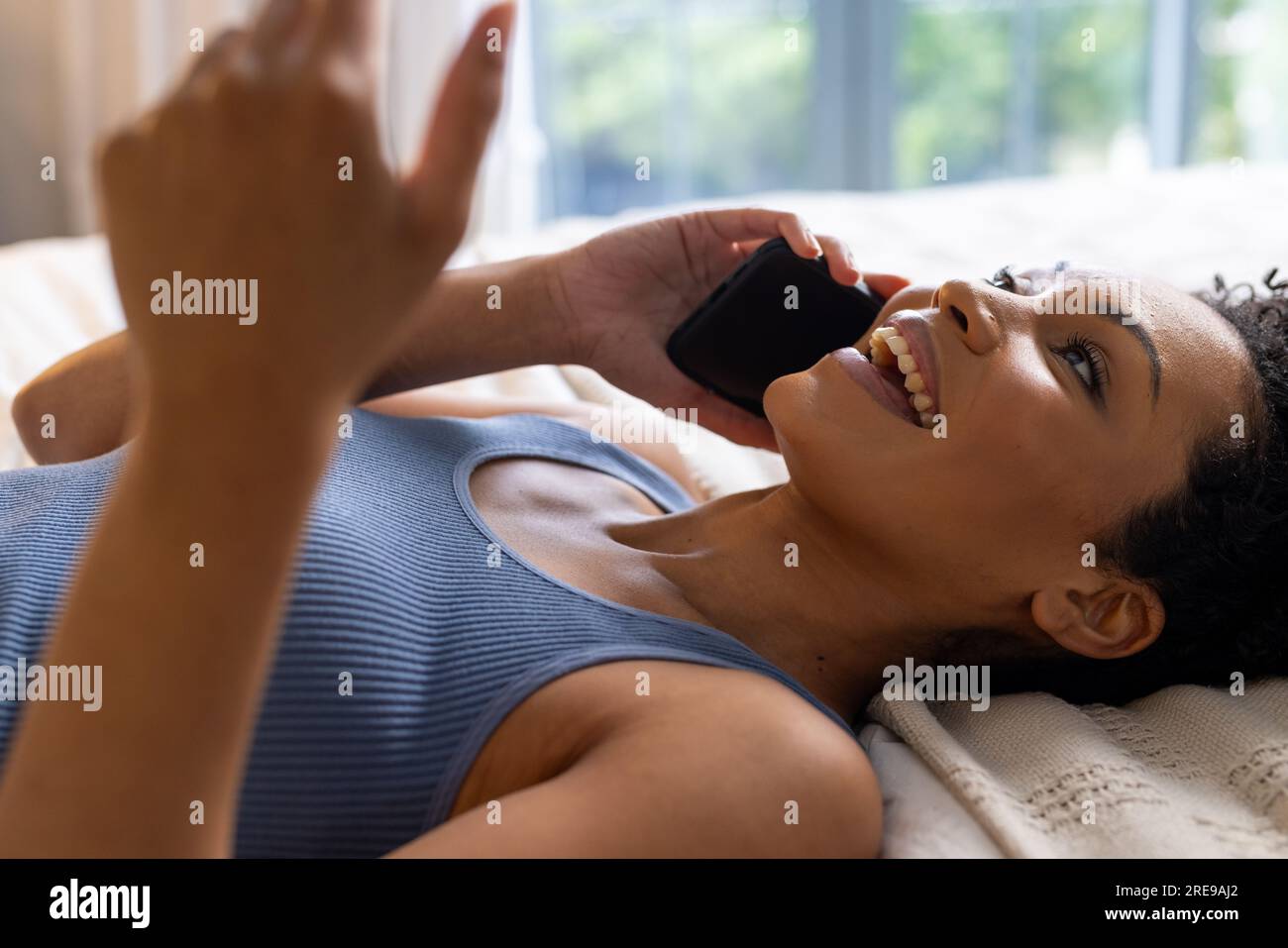 Heureuse femme biraciale allongée sur le lit et parlant sur smartphone dans la chambre Banque D'Images