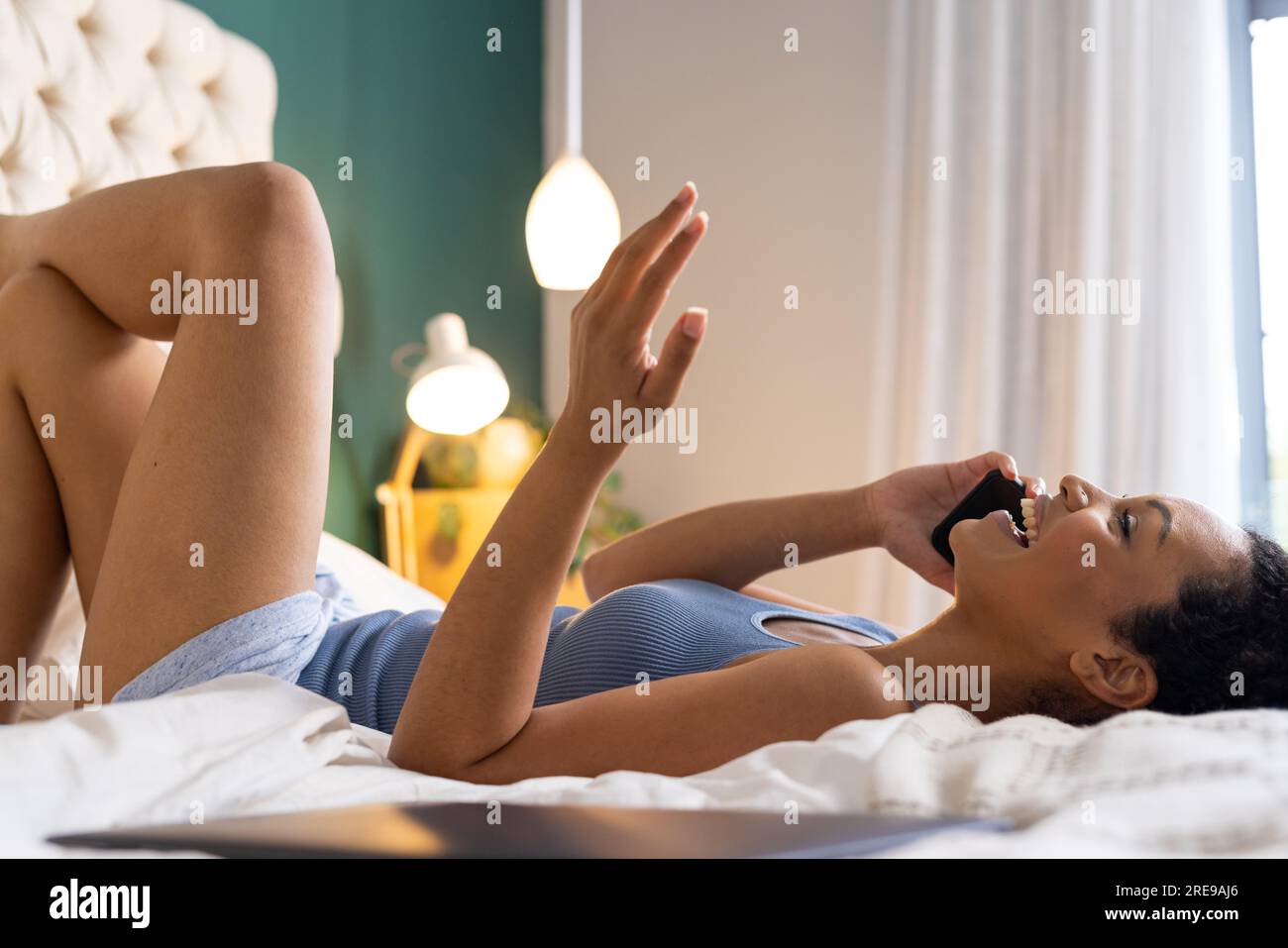 Heureuse femme biraciale allongée sur le lit et parlant sur smartphone dans la chambre Banque D'Images