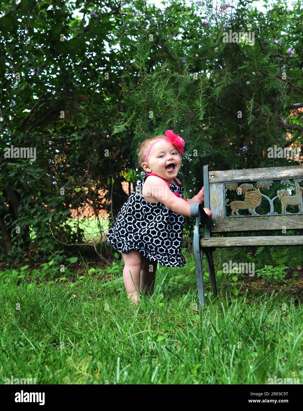 Bébé fille se tient sur un banc en bois et exprime sa joie en gargouillonnant et en ricotant. Elle est pieds nus et porte une robe de robe. Bande de fleur rose encercle Banque D'Images