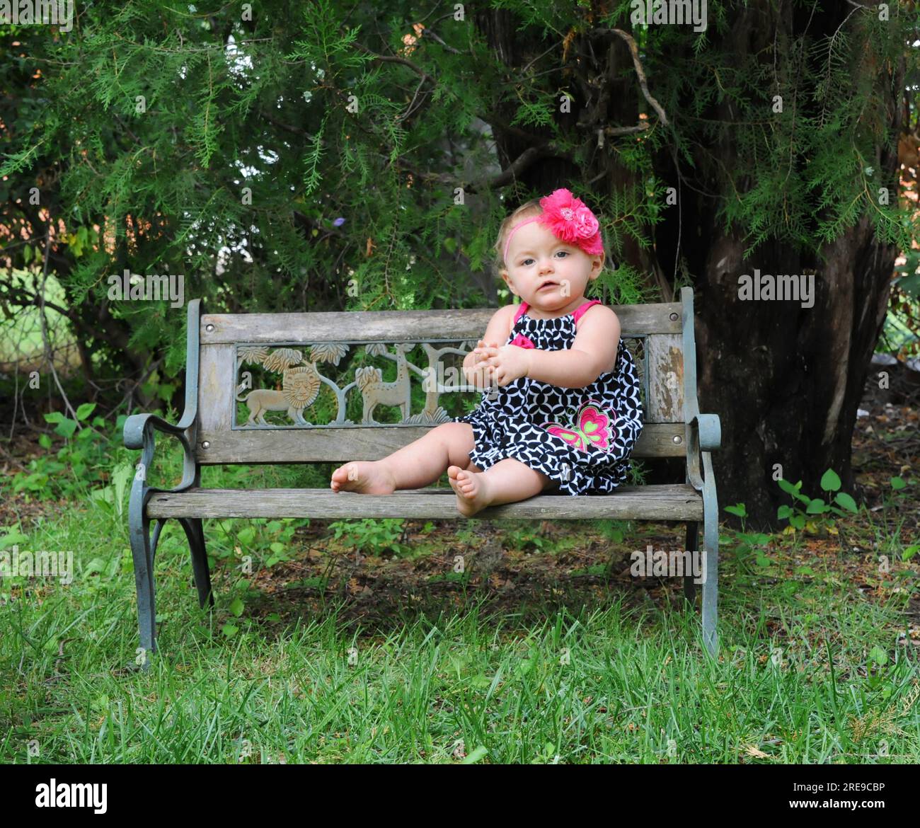 Petite fille assise sur un banc de parc en bois. Elle porte une robe d'été en noir et blanc. Ses cheveux ont un noeud de cheveux de fleur rose. Ses pensées sont quies Banque D'Images