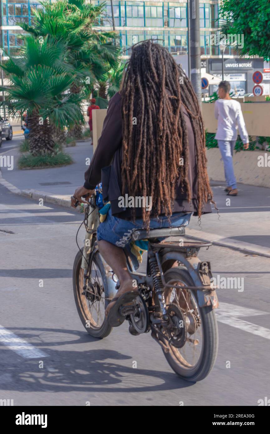 Une photo de derrière d'un homme aux cheveux africains conduisant une vieille moto au milieu de la rue dans une ville Banque D'Images