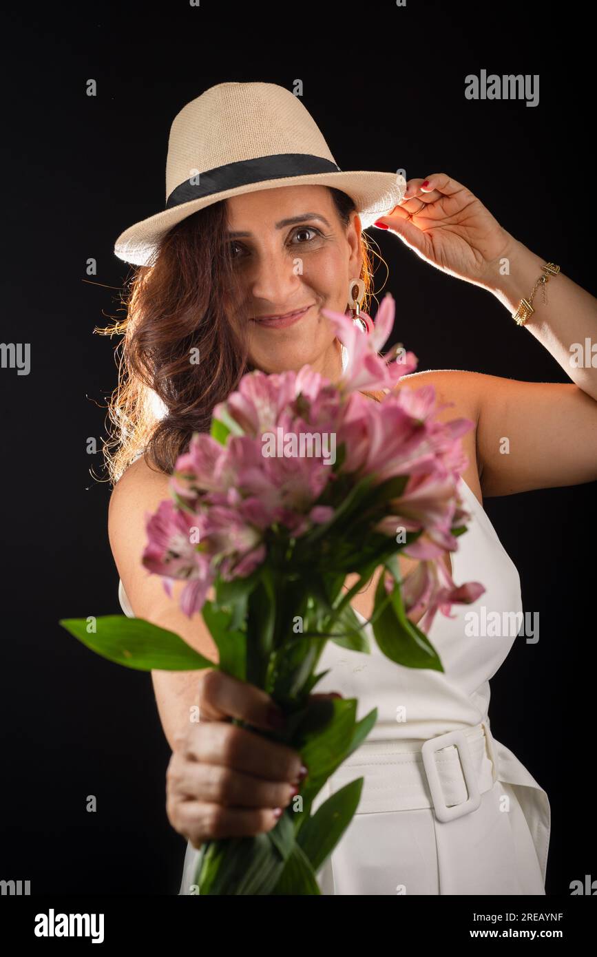 Studio portrait d'une belle femme mûre portant un chapeau tenant un bouquet de fleurs entre les mains. Isolé sur fond noir. Banque D'Images