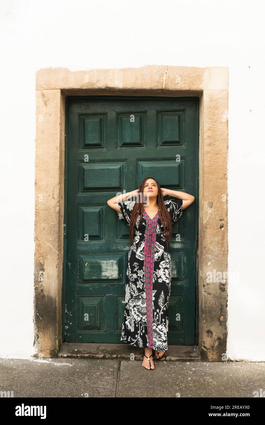Belle femme en tenue colorée avec ses mains derrière sa tête contre une porte de couleur verte. Personne calme et heureuse. Pelourinho, Brésil. Banque D'Images