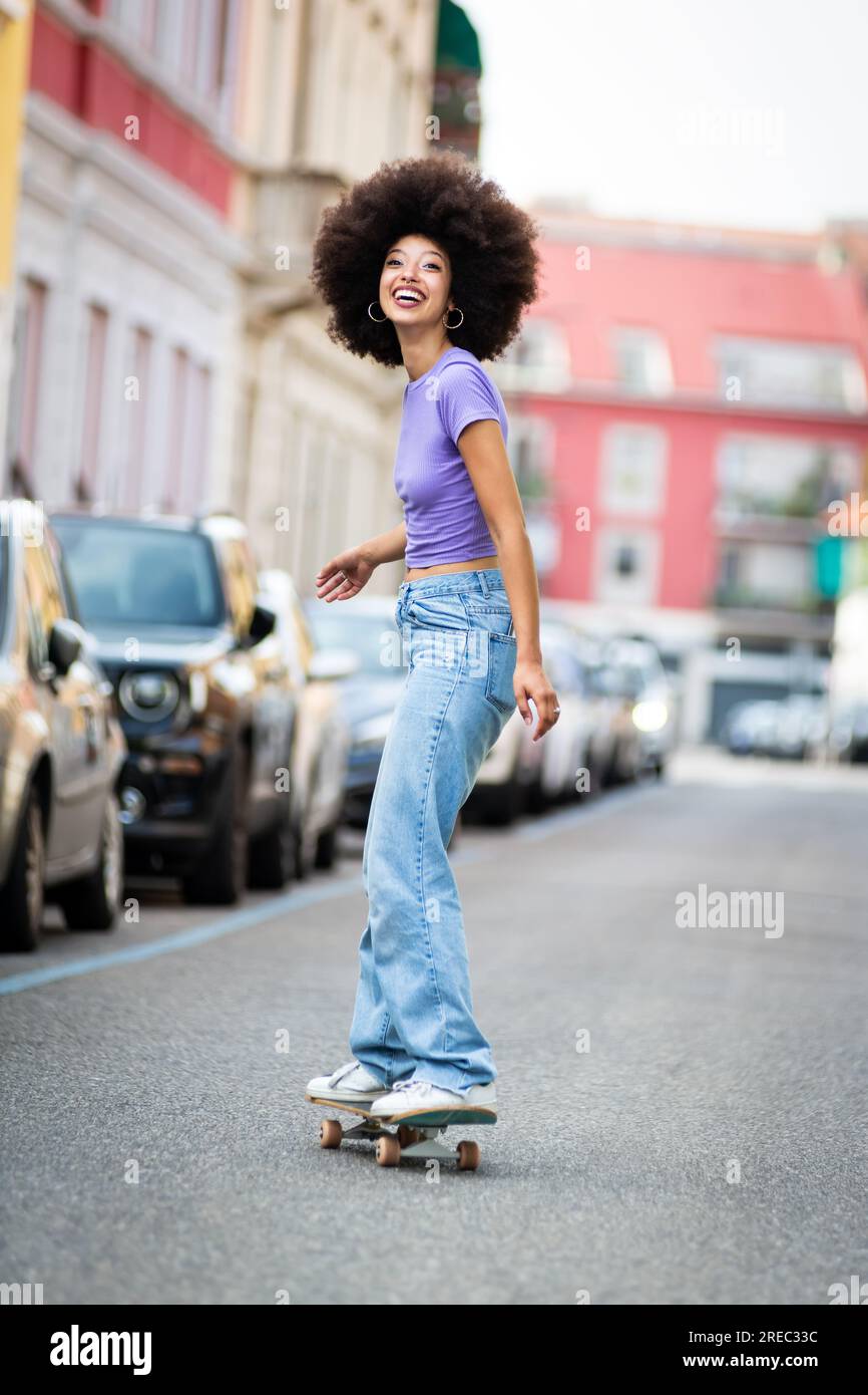 Vue latérale complète du corps de patineuse africaine américaine joyeuse dans des vêtements décontractés avec planche à roulettes afro coiffure sur route asphaltée dans la ville et les toilettes Banque D'Images