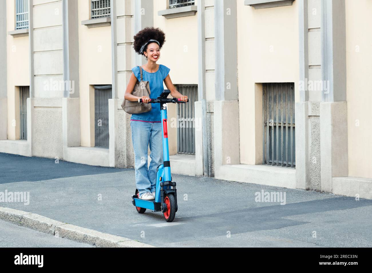 Corps complet de jeune dame noire joyeuse avec coiffure afro chevauchant scooter électrique sur la rue de la ville dans la lumière du soleil Banque D'Images
