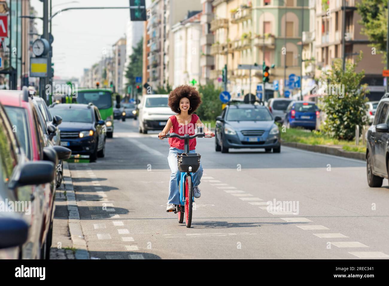 Heureuse femme afro-américaine à vélo sur piste cyclable asphaltée près de la route avec des voitures et regardant la caméra Banque D'Images