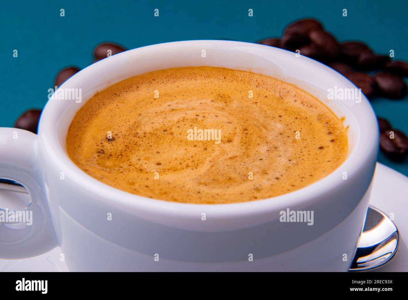 Tasse à expresso blanche et soucoupe avec café brun sur une surface bleu sarcelle avec des grains et une cuillère. Banque D'Images