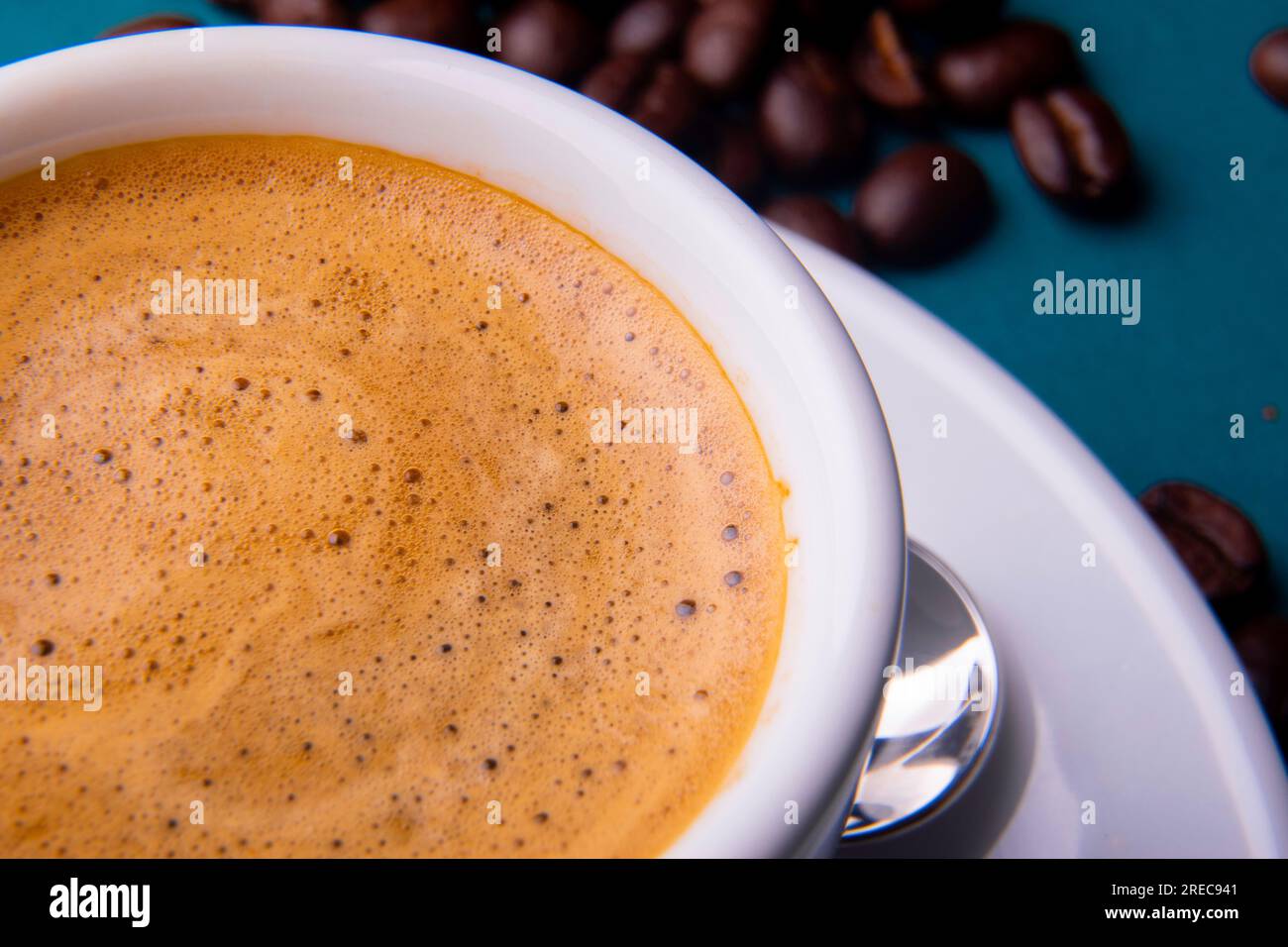 Tasse à expresso blanche et soucoupe avec café brun sur une surface bleu sarcelle avec des grains et une cuillère. Banque D'Images