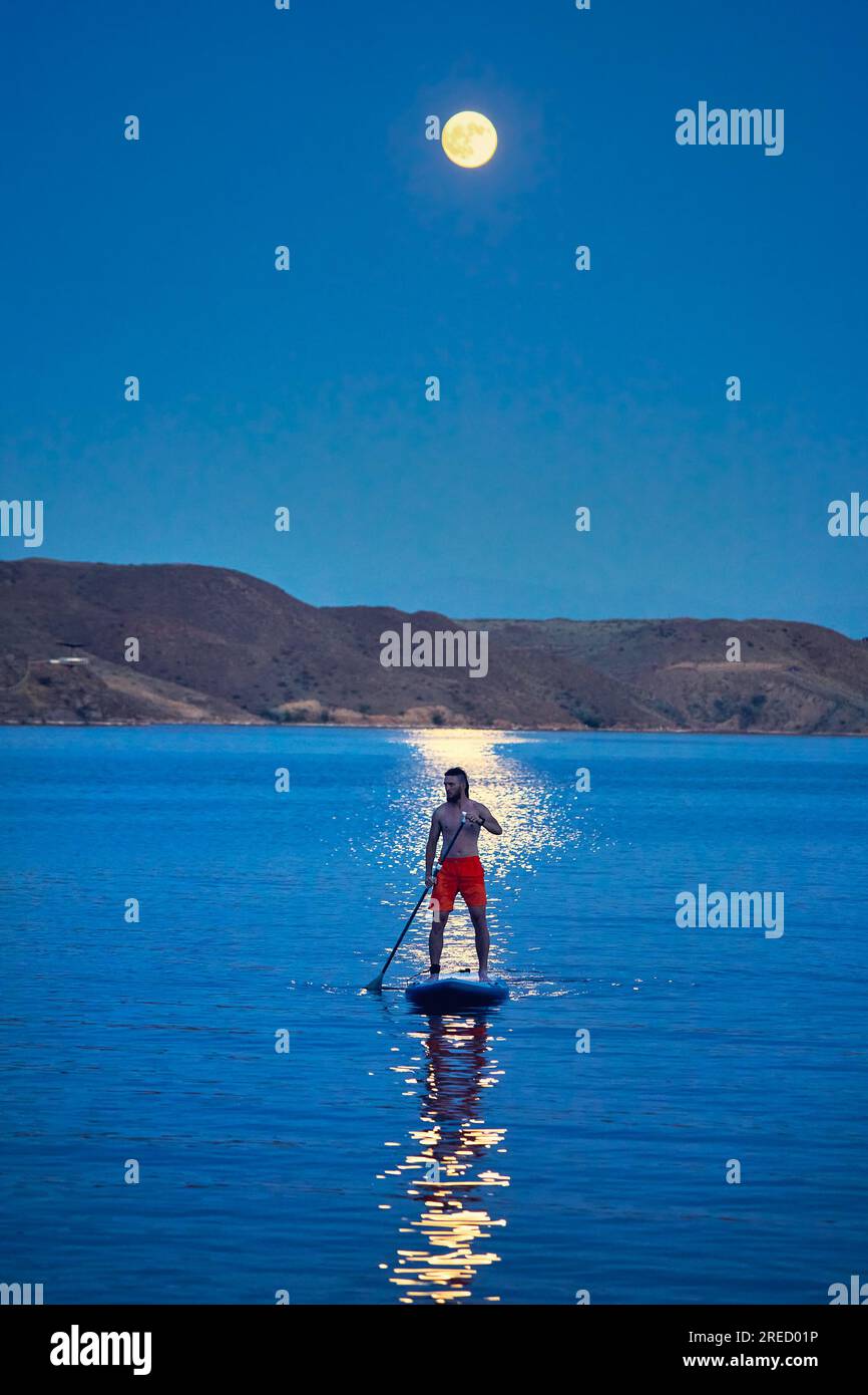 Homme surfeur en chemise orange au stand up paddle boards SUP en silhouette contre la pleine lune chemin lunaire près de l'île au lac Kapchagay au Kazakhstan. Banque D'Images