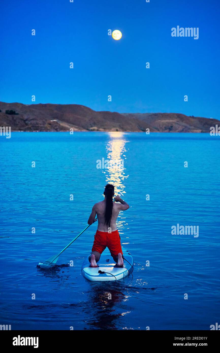Homme surfeur en chemise orange au stand up paddle boards SUP de dos contre la pleine lune chemin lunaire près de l'île au lac Kapchagay au Kazakhstan. Banque D'Images