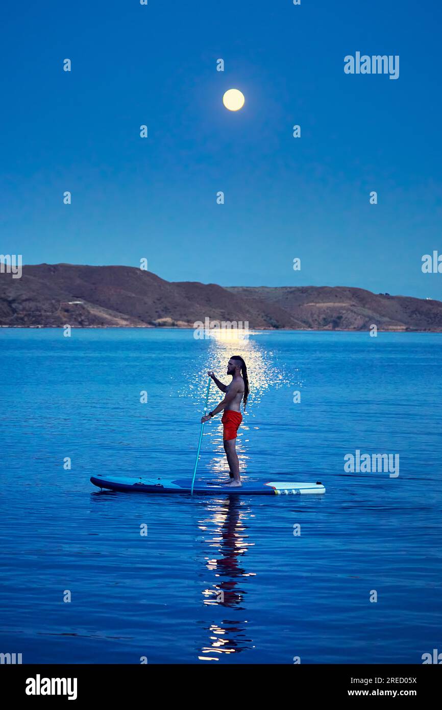 Homme surfeur en chemise orange au stand up paddle boards SUP en silhouette contre la pleine lune chemin lunaire près de l'île au lac Kapchagay au Kazakhstan. Banque D'Images