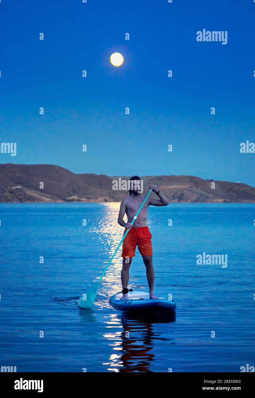 Homme surfeur en chemise orange au stand up paddle boards SUP en silhouette contre la pleine lune chemin lunaire près de l'île au lac Kapchagay au Kazakhstan. Banque D'Images