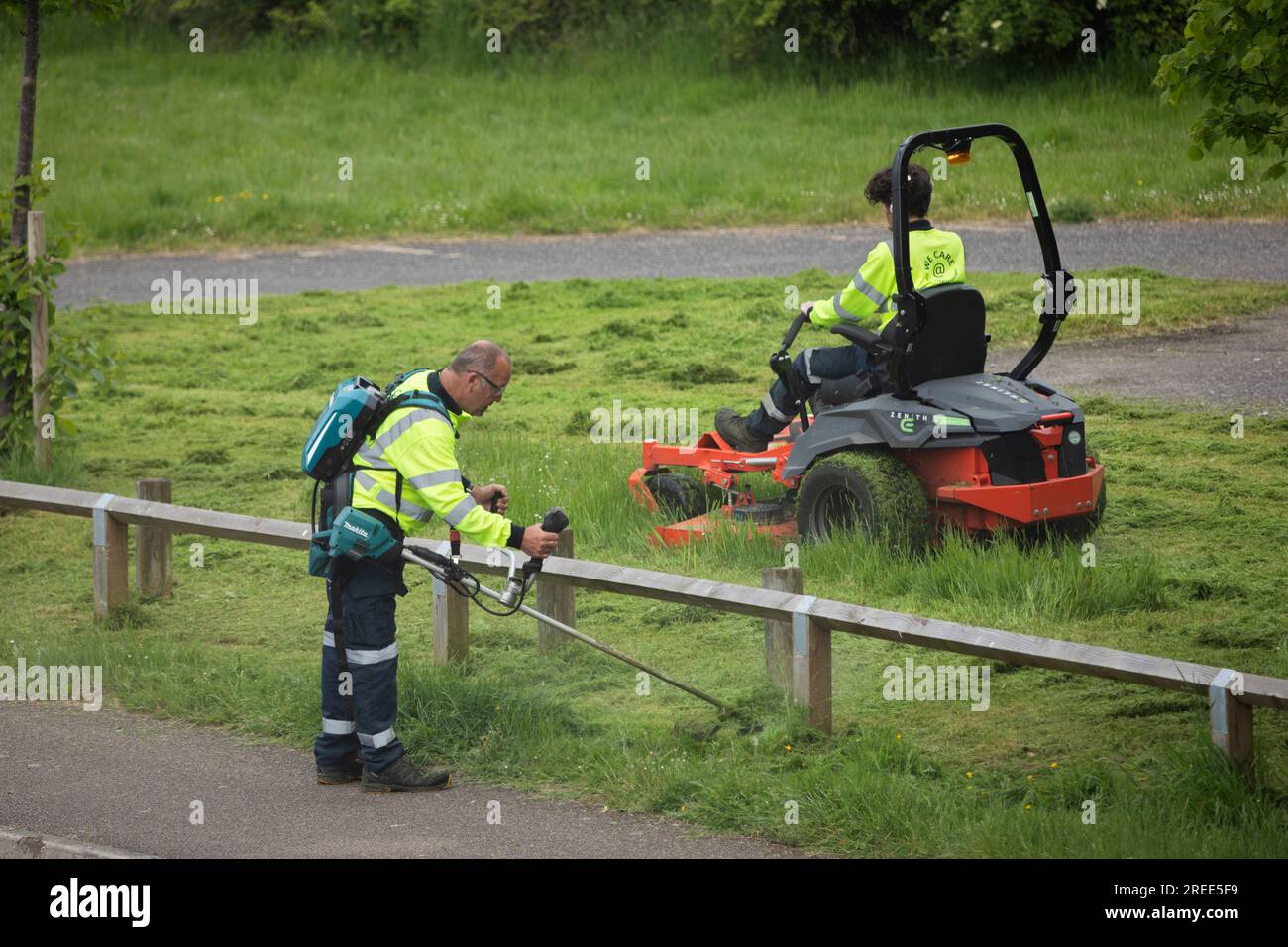 Travailleurs du conseil coupant l'herbe dans l'espace public vu du sentier public, Newbury, Berkshire, Angleterre, Royaume-Uni Banque D'Images