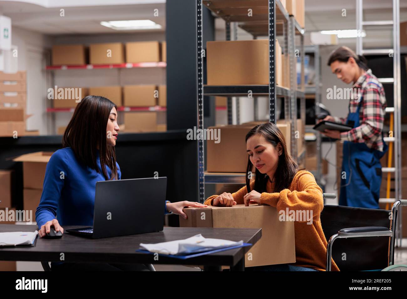 Gestionnaire de livraison d'entrepôt vérifiant la qualité d'emballage de colis tout en travaillant avec le préparateur de commande en fauteuil roulant. Opérateurs d'expédition de magasin préparant la boîte en carton avec des marchandises pour la distribution Banque D'Images