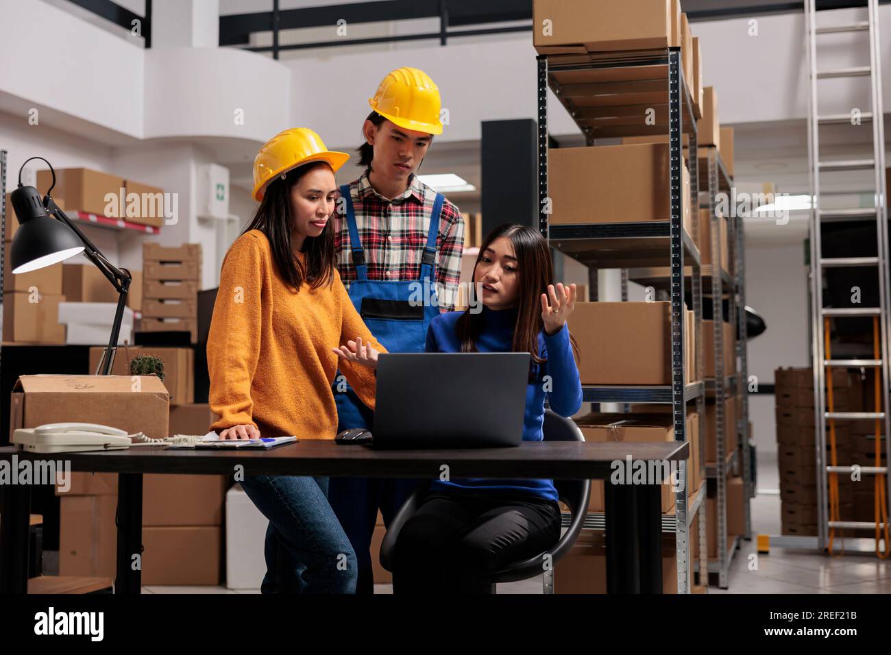 Les travailleurs du centre de distribution asiatique vérifient le calendrier d'approvisionnement des marchandises sur ordinateur portable. Gestionnaire de femme d'entrepôt assis au bureau tout en expliquant la chaîne logistique à des collègues dans la salle de stockage industriel Banque D'Images
