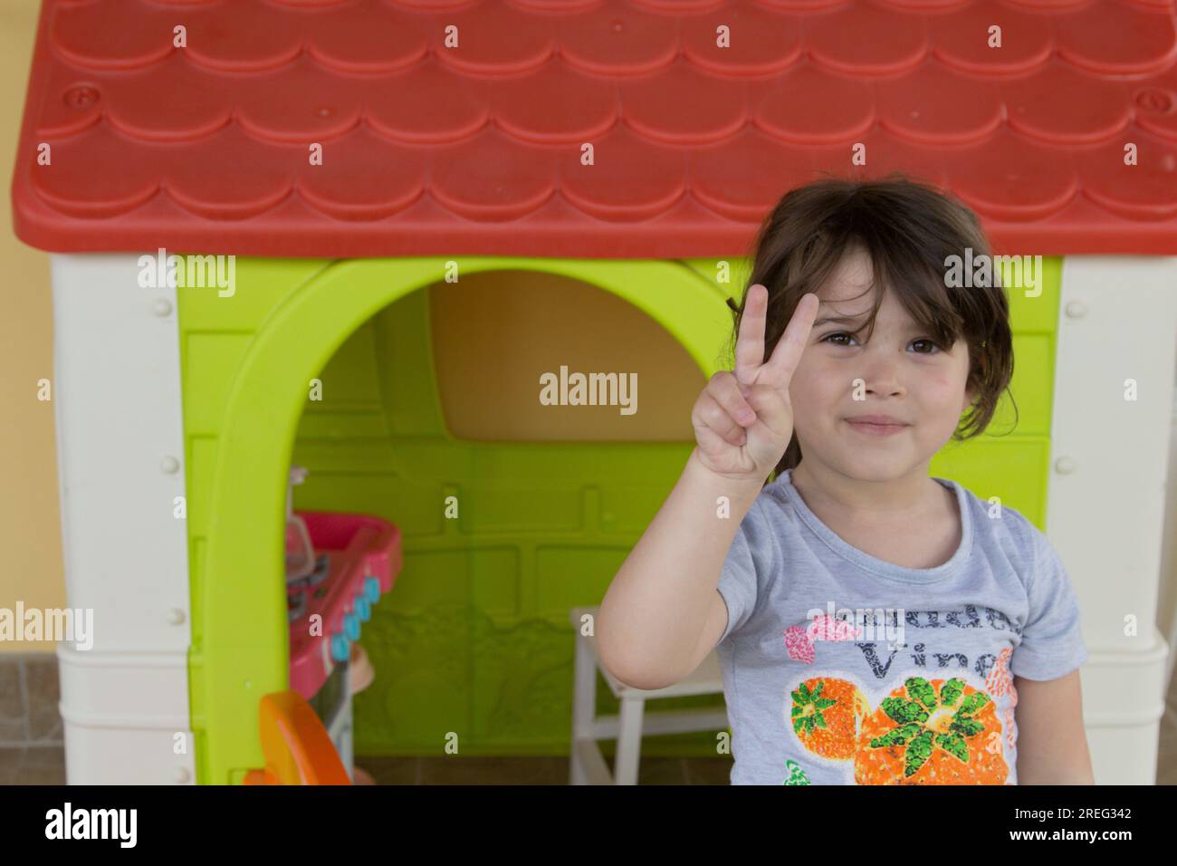 Image d'une adorable petite fille souriante pointant ses doigts vers la victoire tout en jouant dans une maison de jeu dans son jardin. Banque D'Images