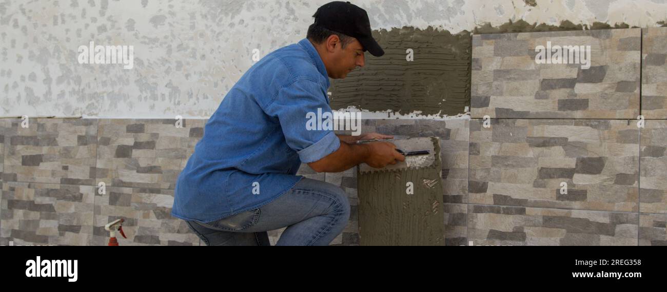 Image d'un carreleur bricoleur qui, avec une spatule, étale la colle pour poser les carreaux sur un mur de maison. Travaux de bricolage et rénovation domiciliaire. Bannière Banque D'Images