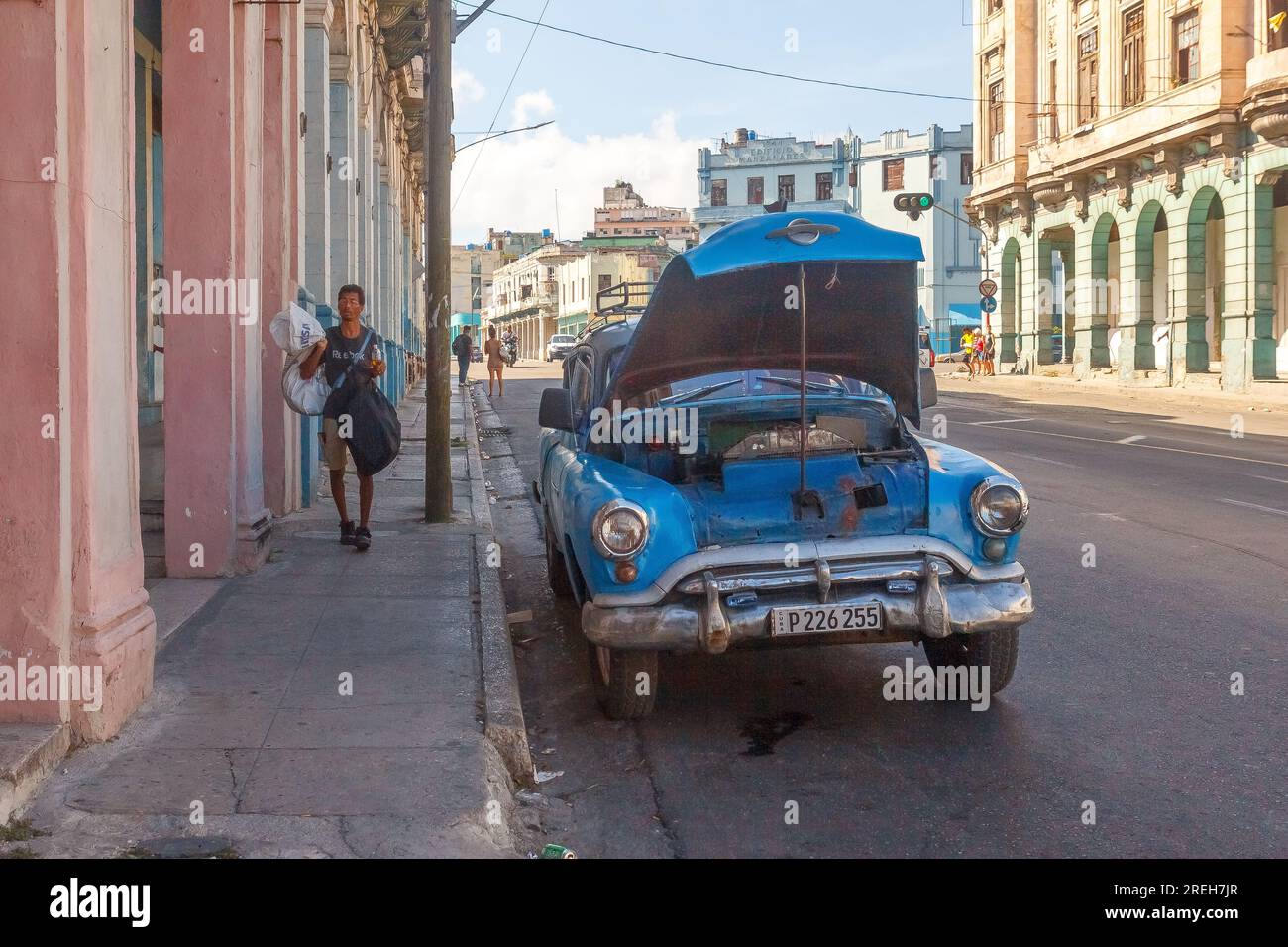 La Havane, Cuba, Une personne cubaine marche sur un trottoir de la ville en portant des sacs. Une voiture Chevrolet vintage cassée a le capot ouvert dans l'avenue. Banque D'Images