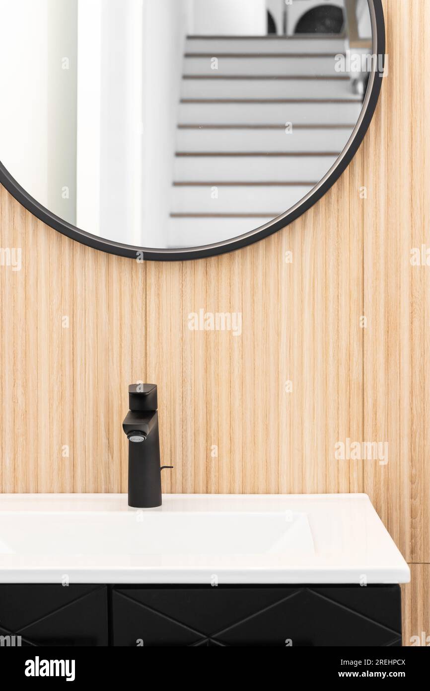 Une salle de bains avec un mur de lattes en bois de chêne blanc avec une armoire noire et, robinet, luminaire, et miroir circulaire. Banque D'Images