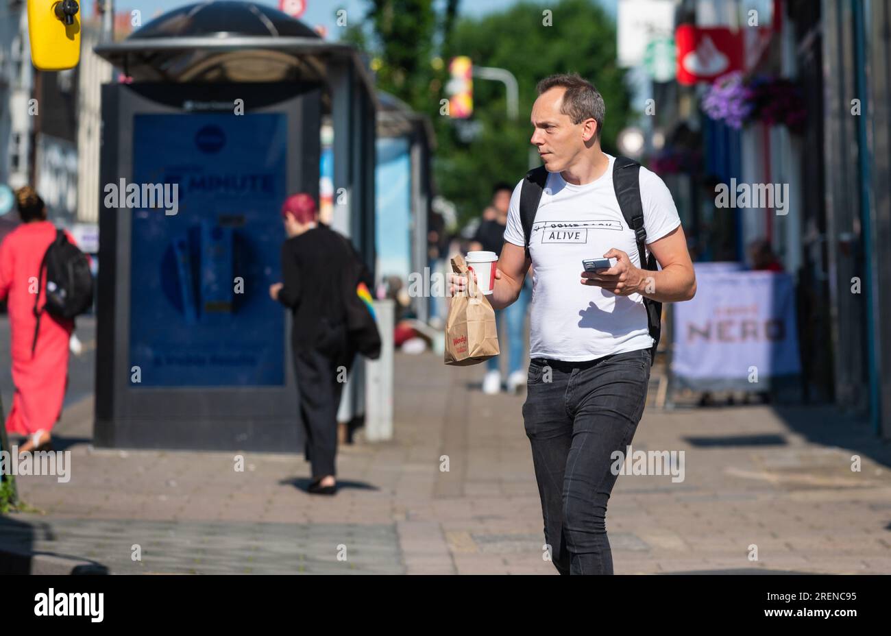 Homme avec sac à dos ou sac à dos, marchant le matin, peut-être marcher au travail, ville occupée, portant téléphone portable, café et petit déjeuner, en été, ROYAUME-UNI. Banque D'Images