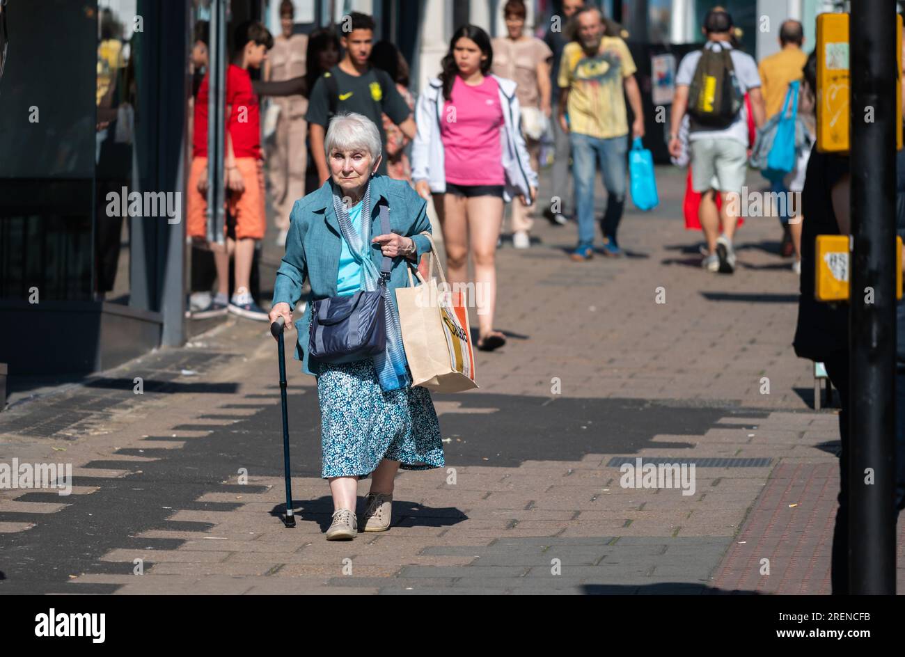 Femme âgée âgée âgée marchant avec bâton de marche ou canne de marche pour l'aide à la marche, dans une rue commerçante animée à Brighton & Hove, Angleterre, Royaume-Uni. Banque D'Images
