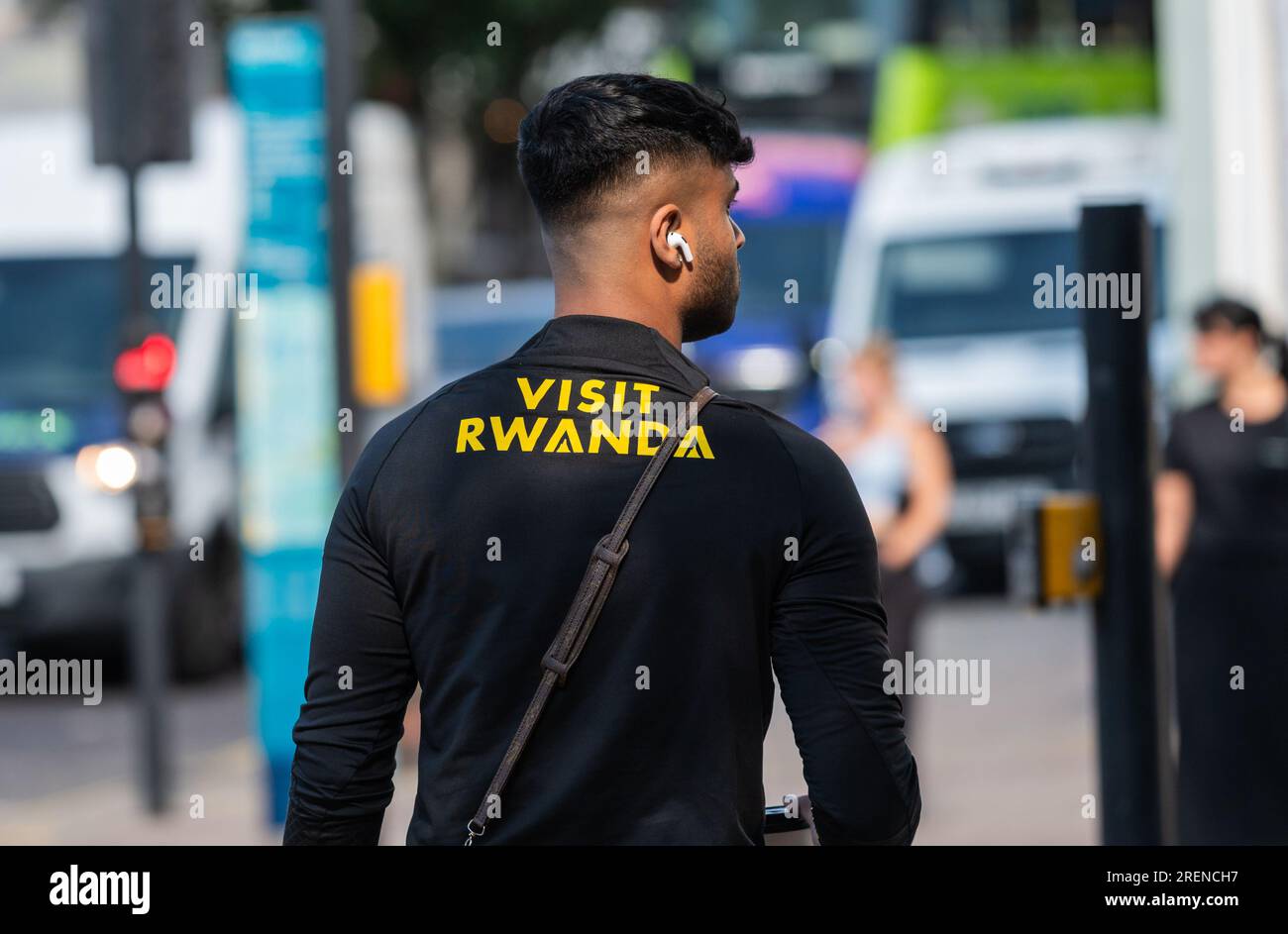 Jeune homme marchant dans une ville portant une chemise Visit Rwanda et portant des écouteurs Bluetooth EarPods ou Airpods sans fil ou une autre marque d'écouteurs intra-auriculaires. Banque D'Images