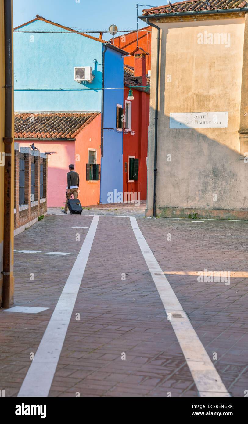 Vue arrière non identifiable d'un voyageur en solo avec un sac de trolley sur les ruelles colorées étroites de Burano, Venise Banque D'Images