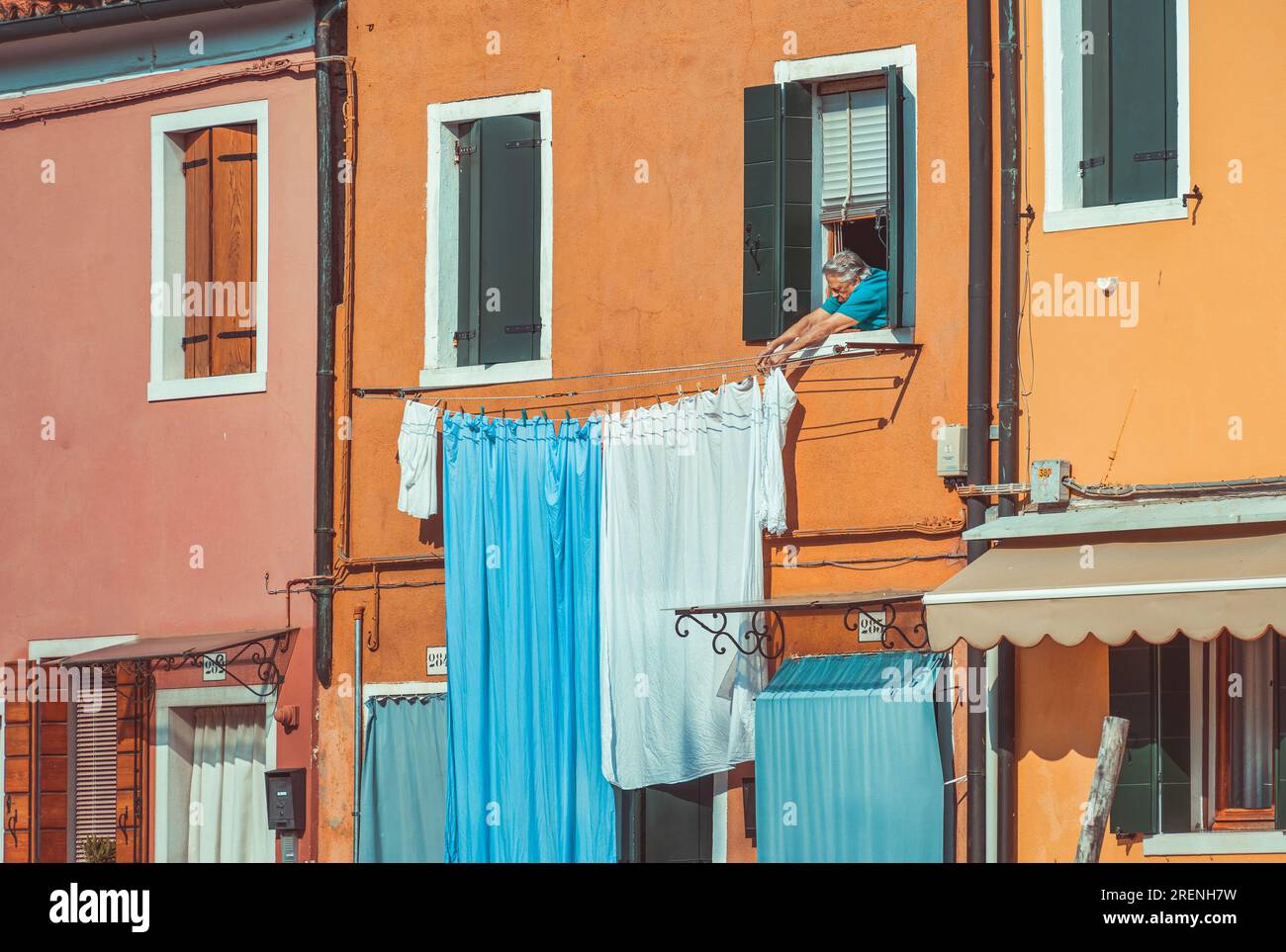 Venise, Italie - Mai 30 2023 : femme locale senior mettant le linge à sécher à la fenêtre. Scène traditionnelle locale de Burano, Italie. Banque D'Images