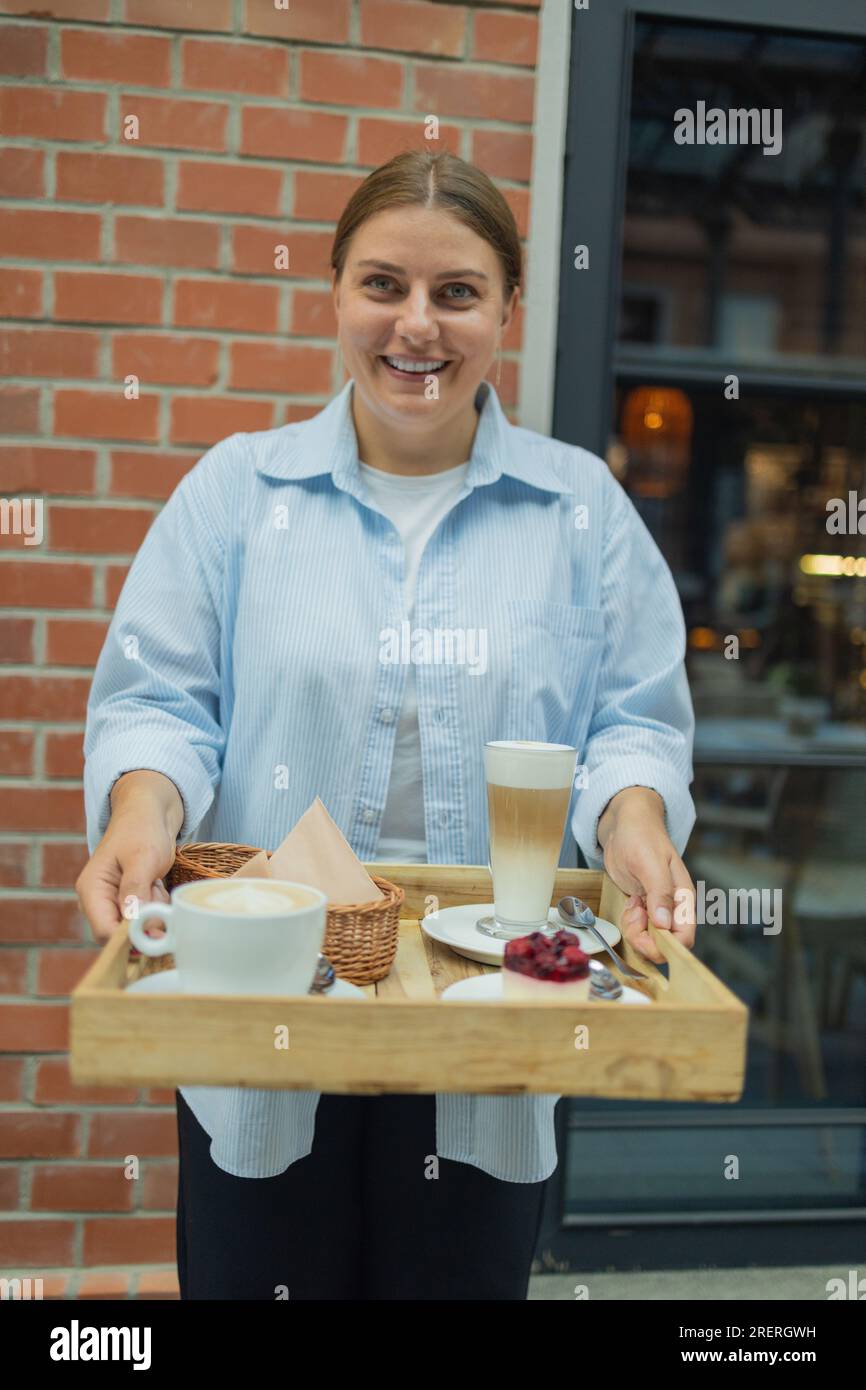 Heureux souriant belle femme serveur dans un restaurant tenant un plateau avec un café latte, cappuccino et gâteau aux baies dans un restaurant Banque D'Images