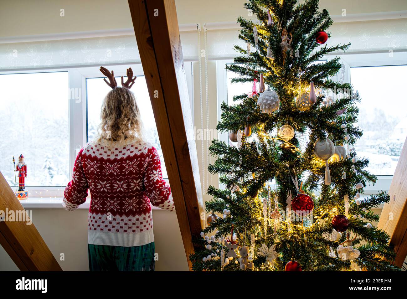 Femme adulte méconnaissable portant un pull de Noël à motif rouge regardant par la fenêtre de la maison et attendant les vacances de Noël à côté de l'arbre de Noël. Banque D'Images