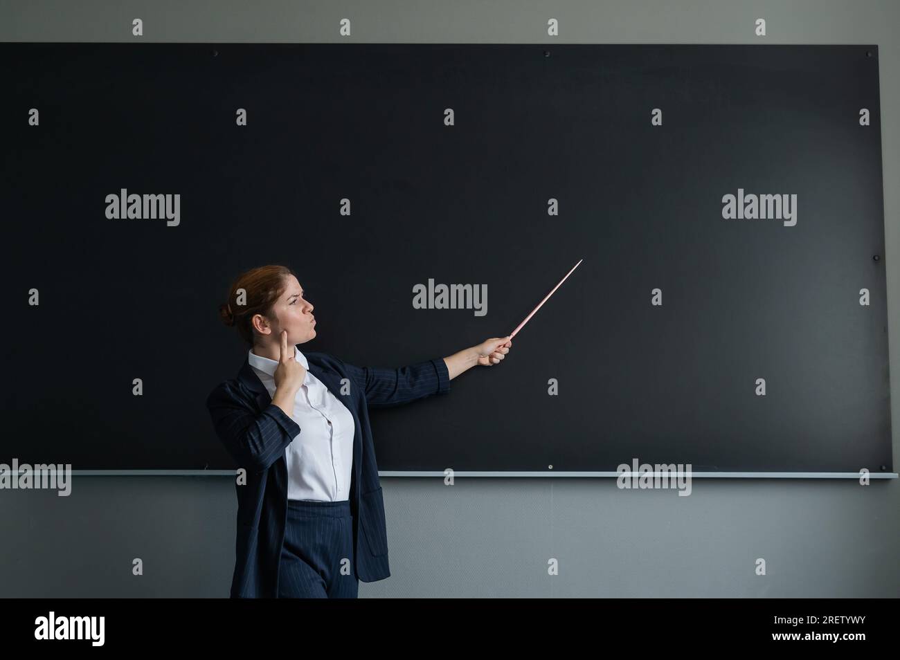 Femme caucasienne aux cheveux rouges dans un costume de pantalon. Enseignante réfléchie avec un pointeur au tableau noir. Banque D'Images