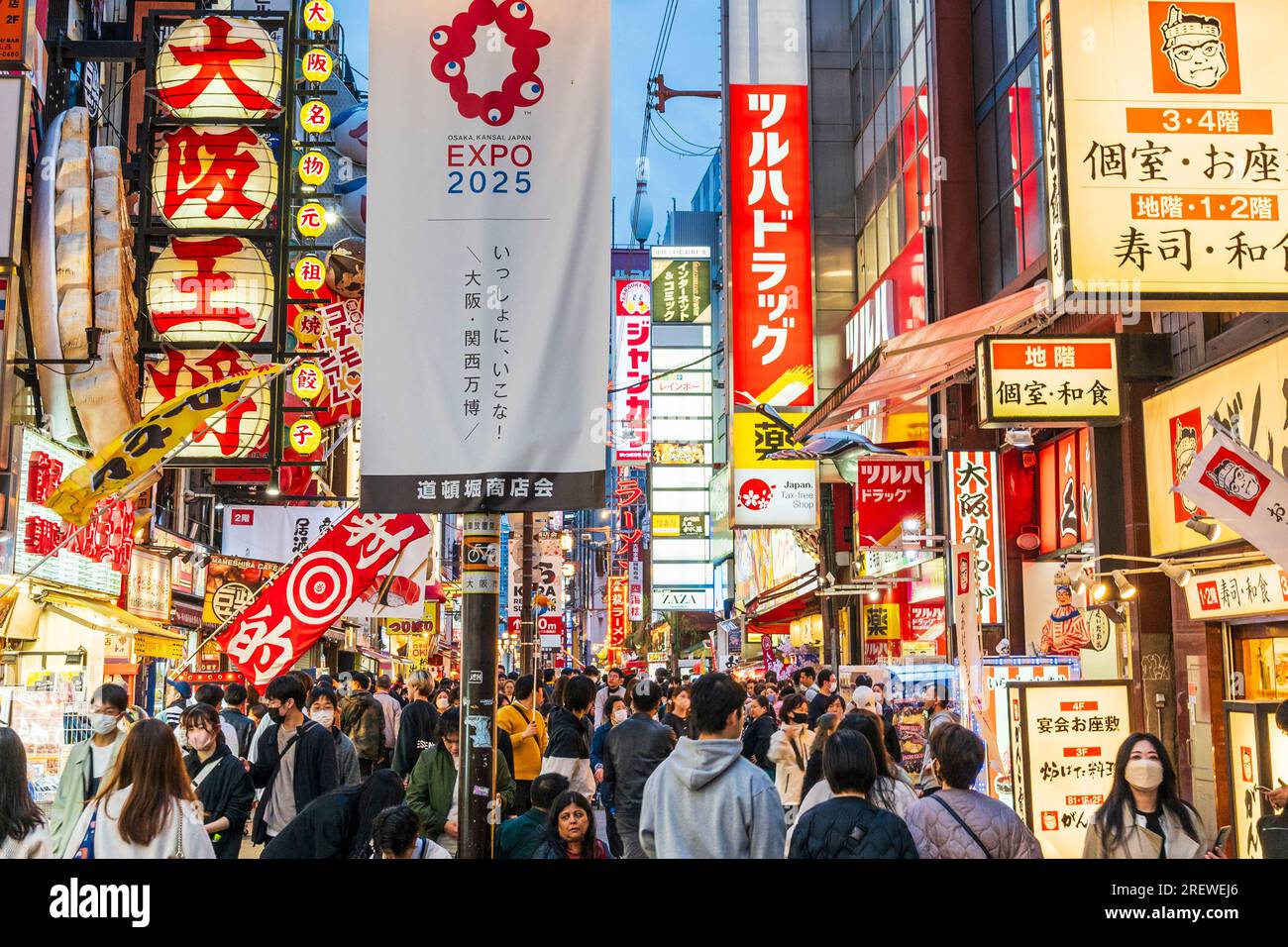 Vue le long de Dotonbori, début de soirée, occupé avec les gens. Stand d'éclairage de rue de premier plan avec bannière expo 2025, derrière que de nombreux panneaux au néon. Banque D'Images