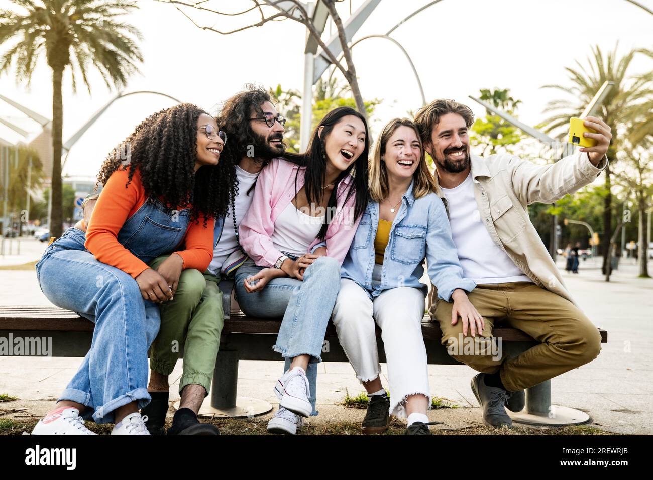 Groupe diversifié d'amis prenant un selfie dans la rue assis sur un banc. Joyeux groupe multiracial de jeunes hipsters prenant une photo à l'extérieur dans un Banque D'Images