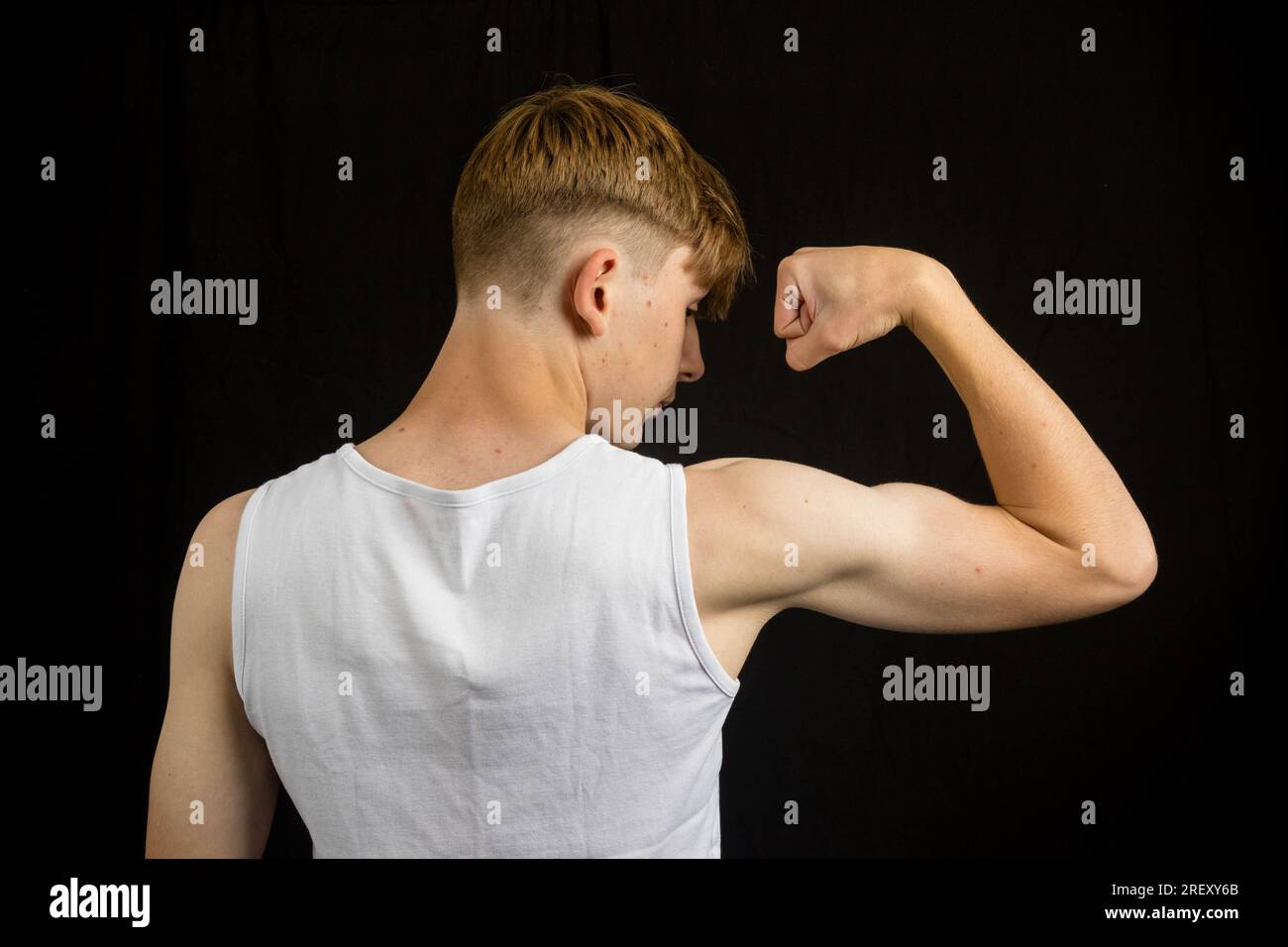 Portrait d'un adolescent caucasien de 14 ans portant un gilet sans manches fléchissant son biceps par derrière contre un garçon de fond noir Banque D'Images