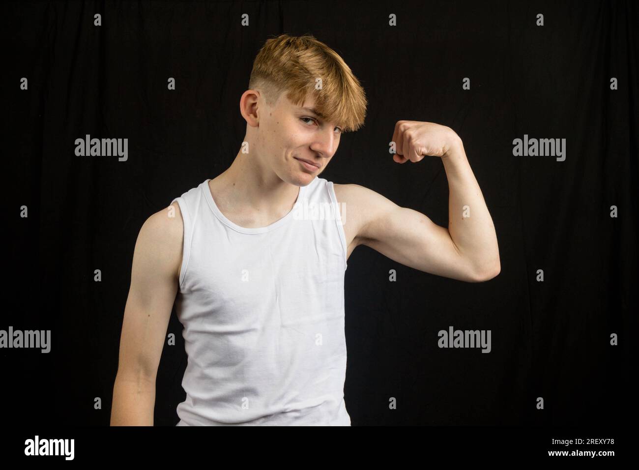 Portrait d'un adolescent caucasien de 14 ans portant un gilet sans manches fléchissant son biceps sur un fond noir Banque D'Images