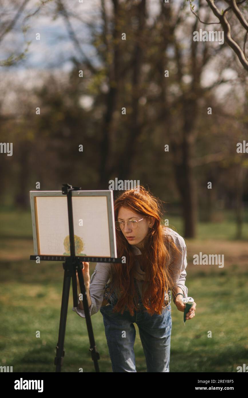 Femme de gingembre debout à côté d'un chevalet et peinture sur elle dans la nature au parc Banque D'Images