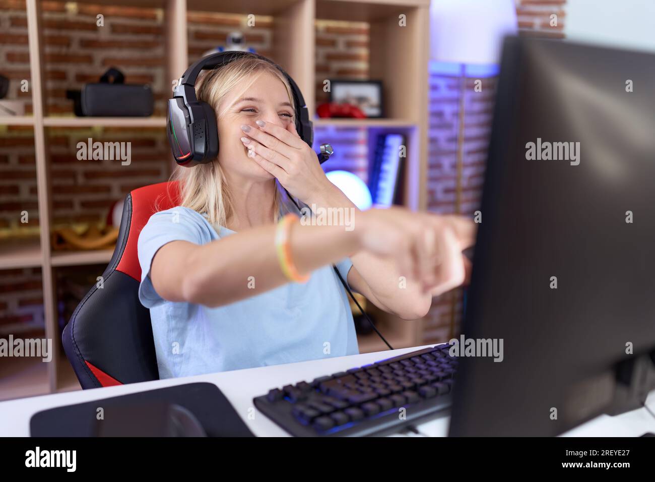 Jeune femme caucasienne jouant à des jeux vidéo portant des écouteurs riant de vous, pointant du doigt la caméra avec la main sur la bouche, expression de honte Banque D'Images