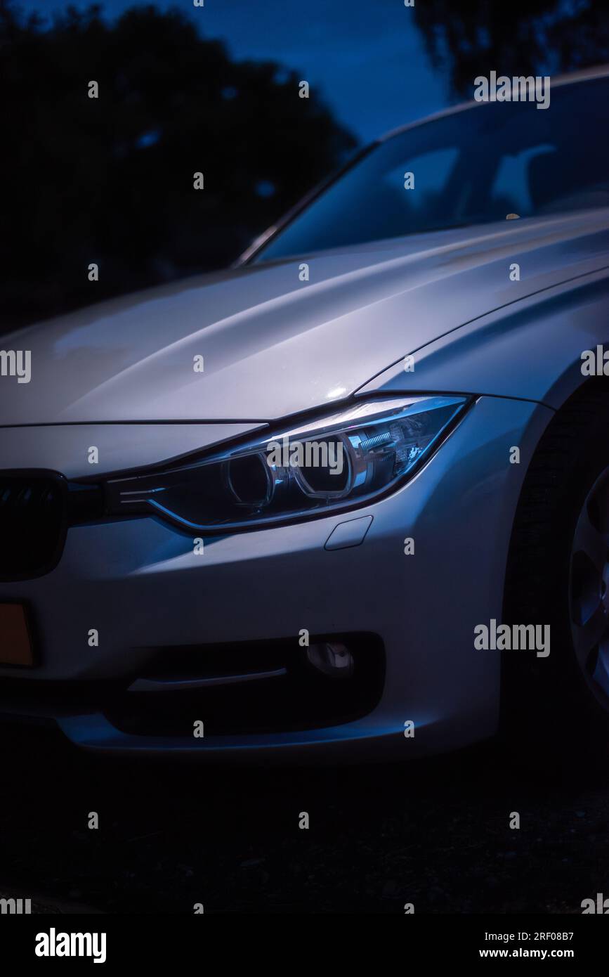 Detailshot d'un phare BMW série 3 de 2012. Pris la nuit, un lampadaire met en valeur le capot et le phare de la voiture argentée. Banque D'Images