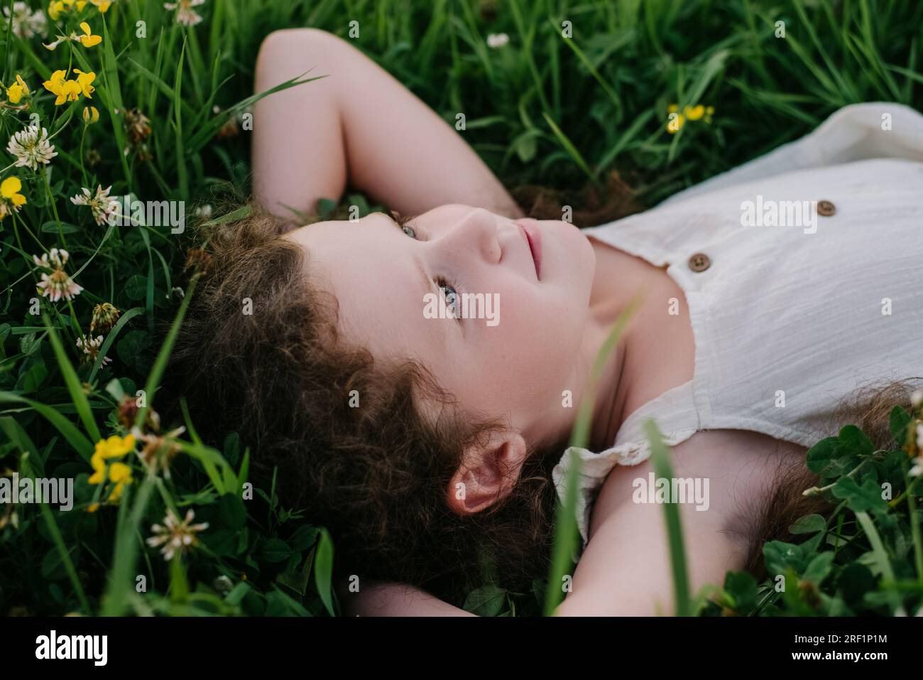 Vue de côté de heureux joli petit enfant de fille couché sur l'herbe verte dans la journée chaude d'été. Portrait enfant adorable rêve en regardant le ciel sur la pelouse à l'extérieur Banque D'Images