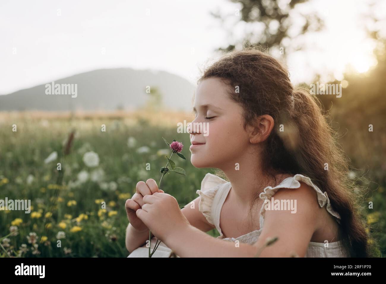 Portrait de souriant charmante petite fille en robe aimez sentir les fleurs sauvages assis dans la prairie à l'extérieur pendant le coucher du soleil chaud. Saison estivale. Childho Banque D'Images